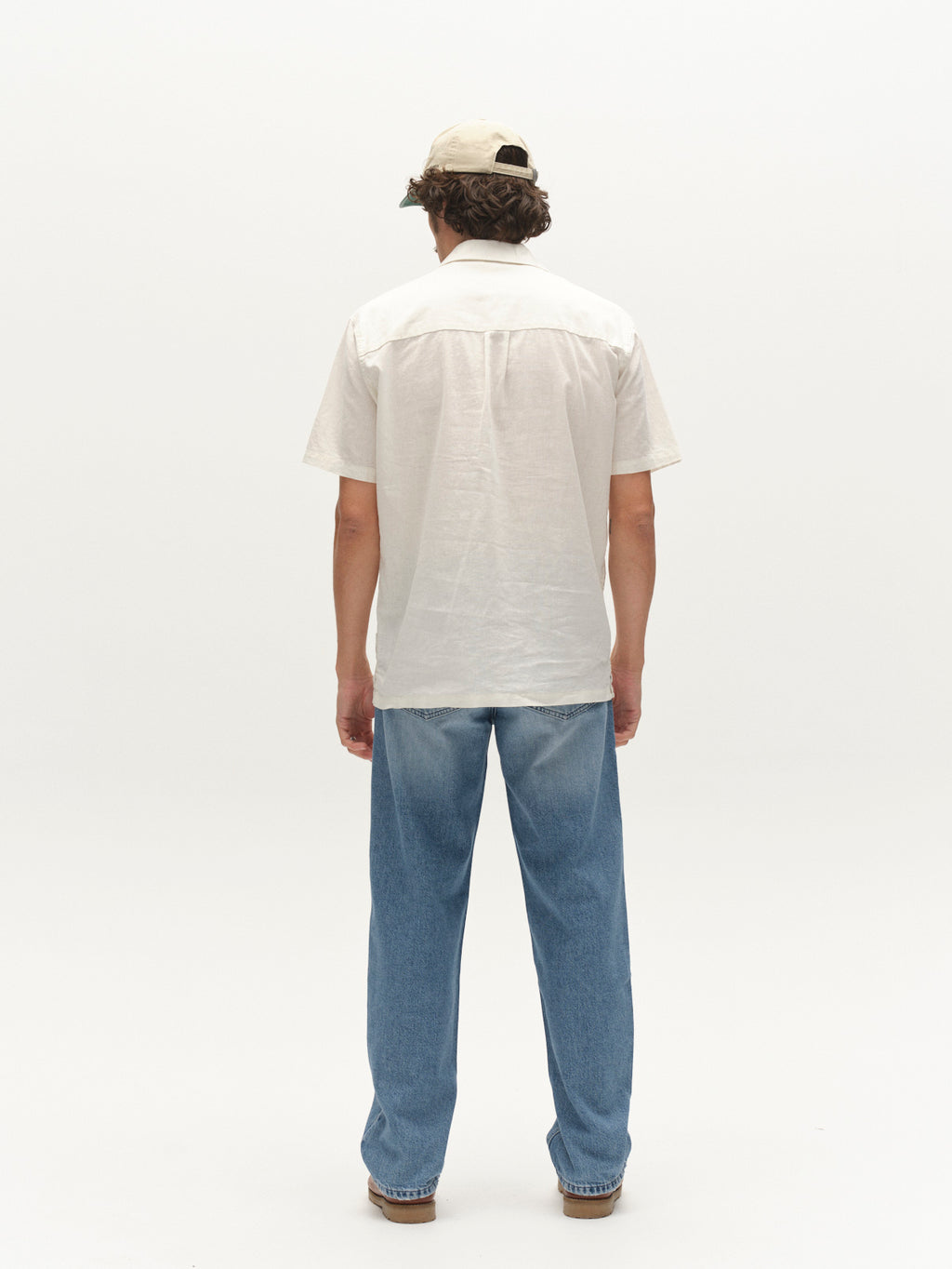 A person faces away from the camera, wearing the Gabba GABtimo Resort Lins SS Shirt in Off White, light blue jeans, brown shoes, and a beige cap against a plain white background.