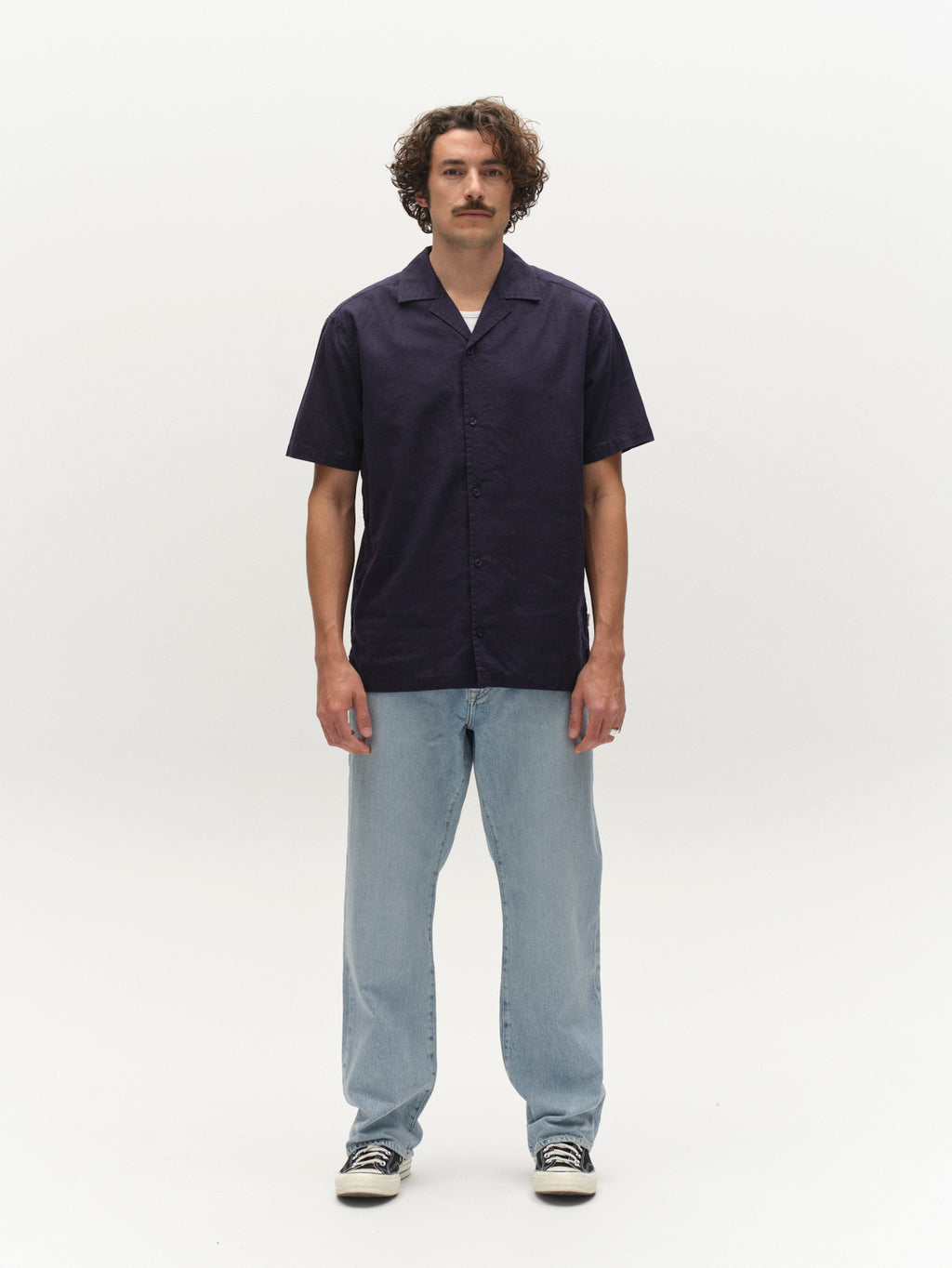 A person with curly hair and a mustache faces forward against a plain white background, wearing the Gabba GABtimo Resort Lins SS Shirt in Navy, light blue jeans, and black-and-white sneakers.