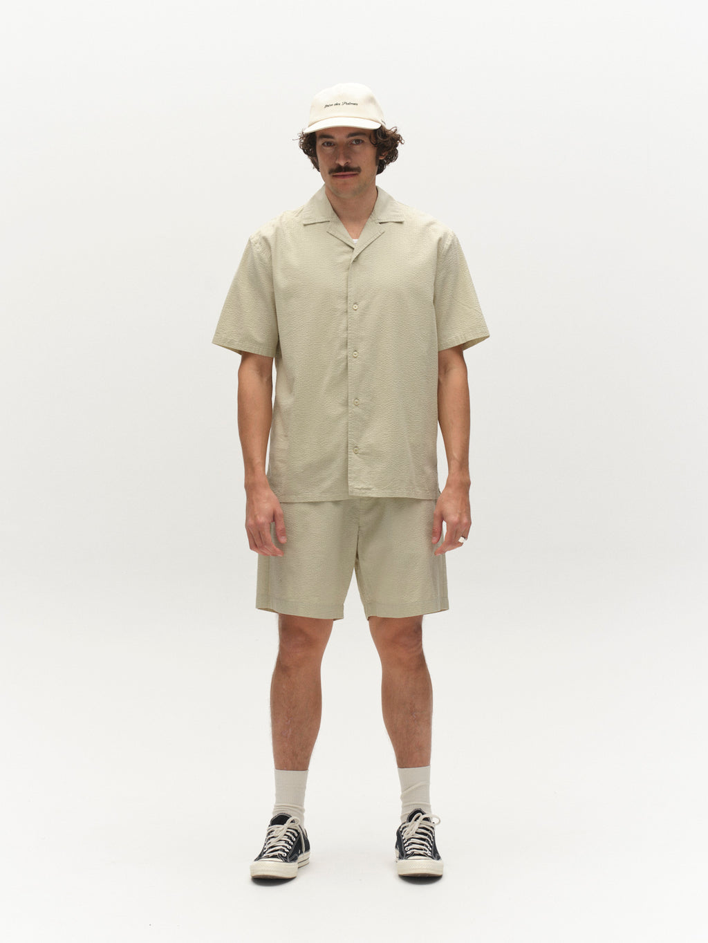 A man with a mustache stands against a white background, wearing the Gabba GABtimo Resort Ker SS Shirt in Alfa Green. He pairs it with matching shorts, a beige bucket hat, white socks, and black sneakers.