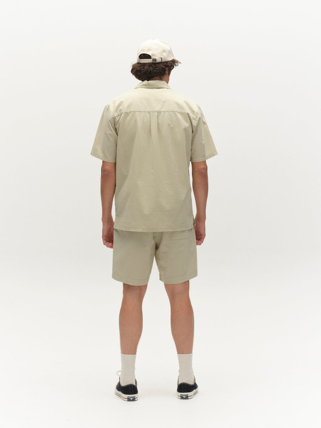 A person stands facing away, wearing the Gabba GABtimo Resort Ker SS Shirt in Alfa Green with matching shorts, a beige cap, white crew socks, and black shoes against a plain white background.