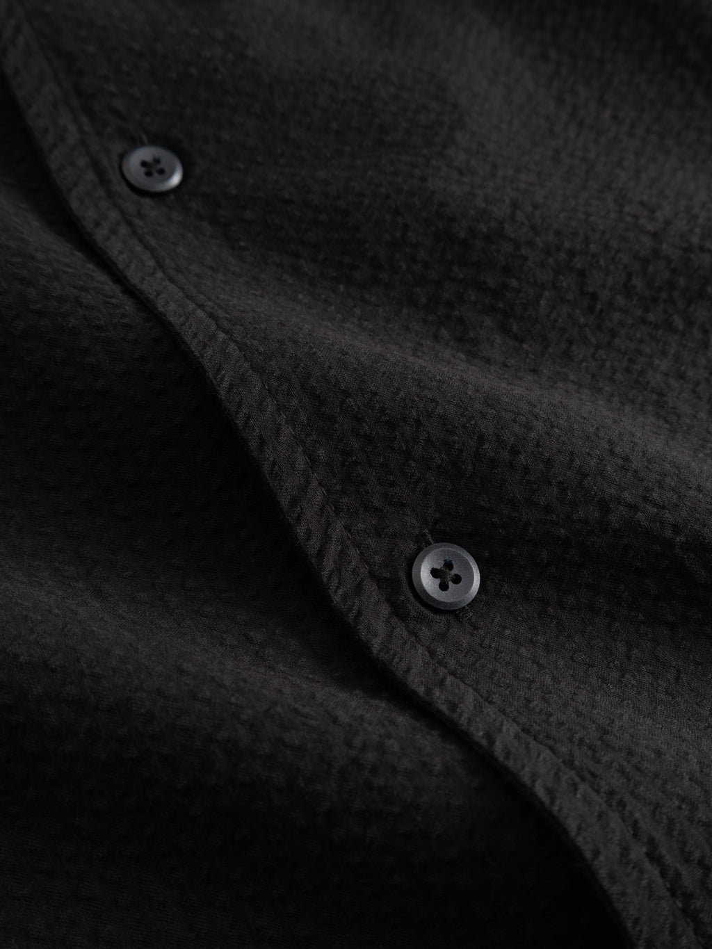 Close-up of the Gabba GABtimo Resort Ker SS Shirt in black cotton seersucker, featuring two black buttons on a seam, a subtle pattern, textured stitching, and short sleeves.