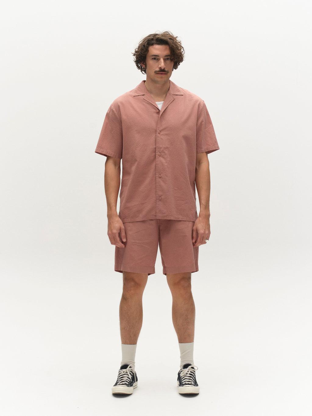 A man with curly hair and a mustache stands facing forward, wearing a pink short-sleeve button-up shirt, Gabba GABtommi Ker Shorts in Wood Pink, white socks, and black-and-white sneakers against a plain white background.