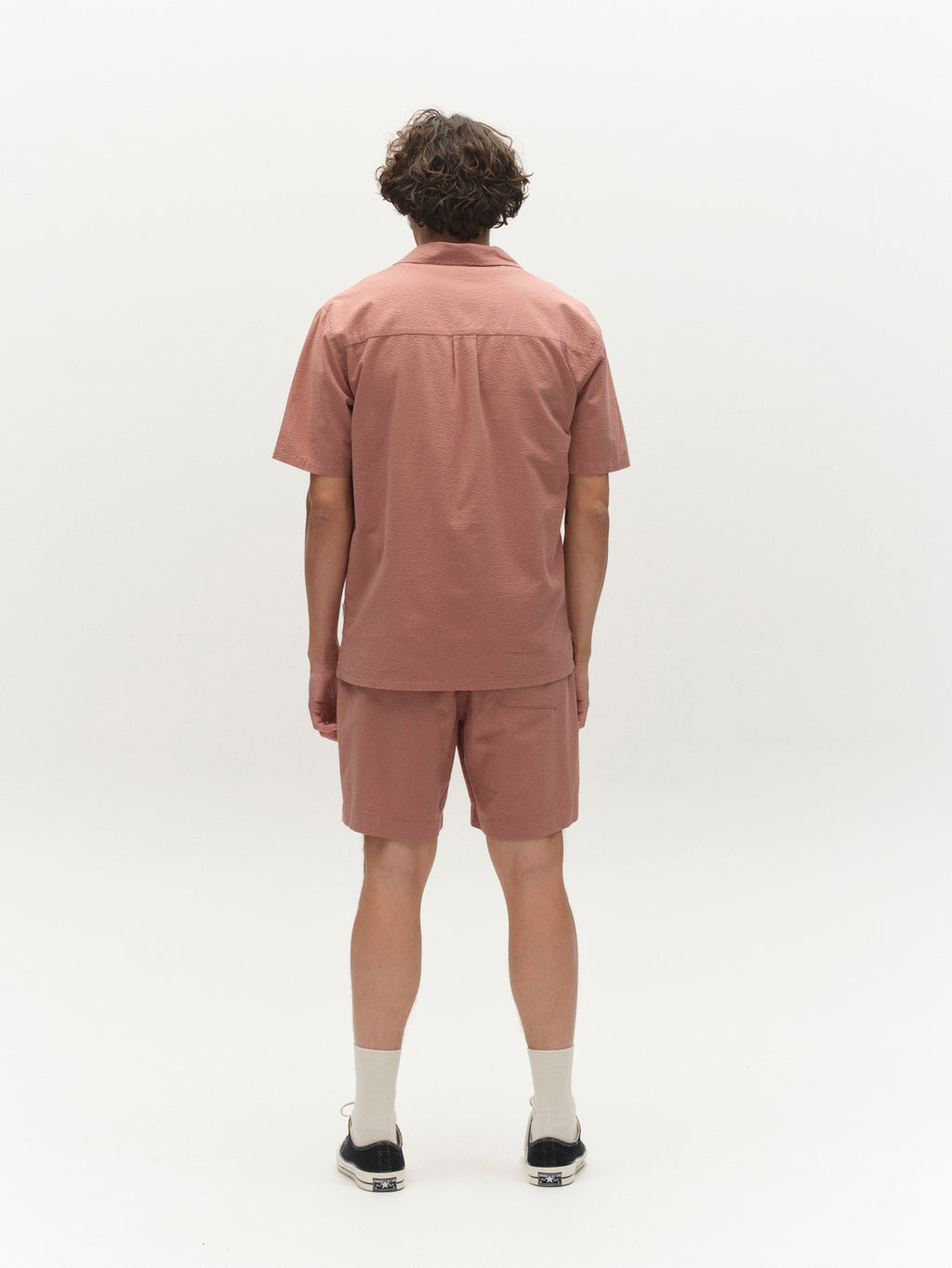 A person with short, curly hair is seen from behind in a light pink short-sleeve shirt and Gabba GABtommi Ker Shorts - Wood Pink with an elastic waistband, paired with white socks and black shoes against a plain white background.