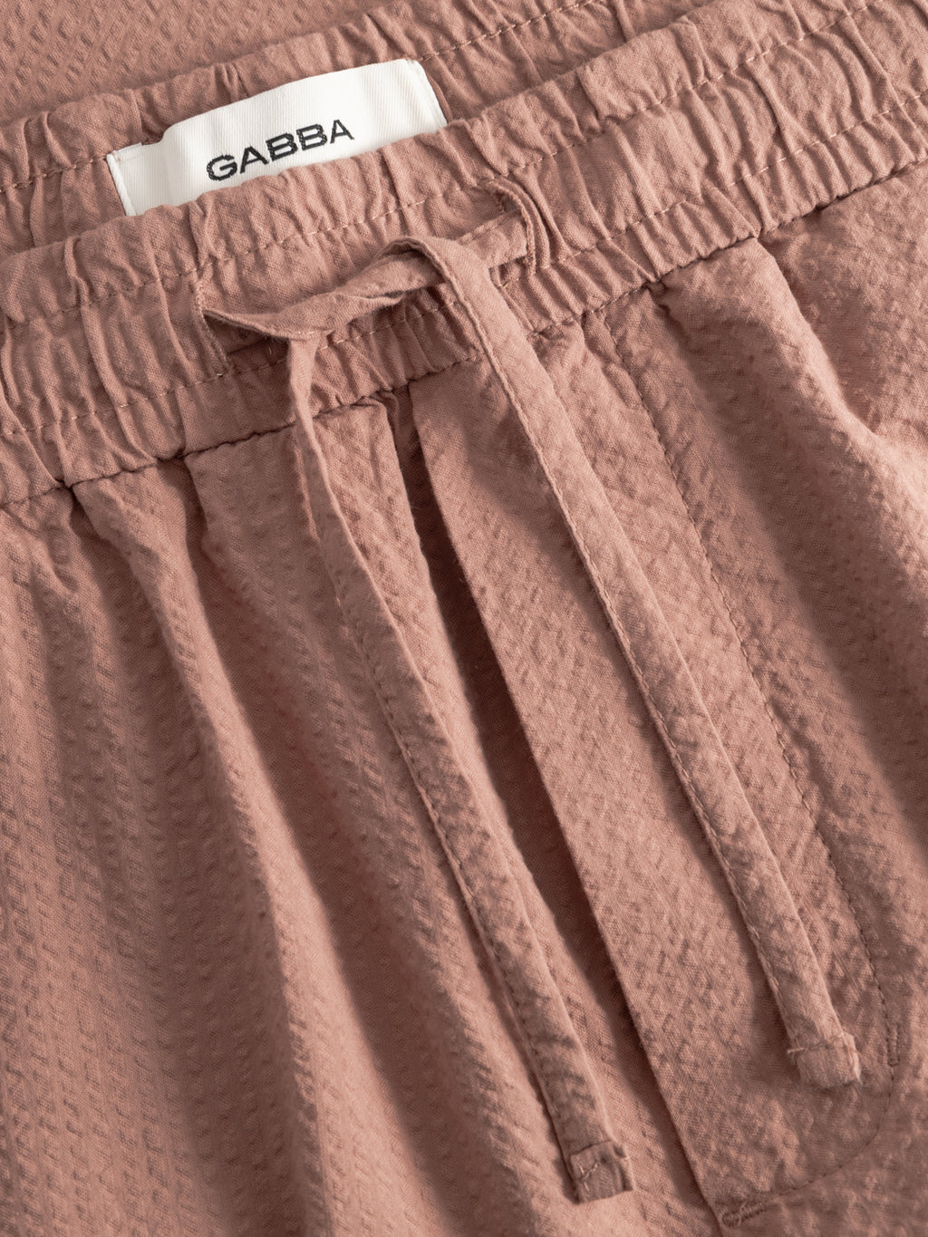 Close-up of textured Wood Pink GABtommi Ker Shorts by Gabba, featuring an elastic waistband, adjustable drawstring, and a white label with the Gabba brand name.