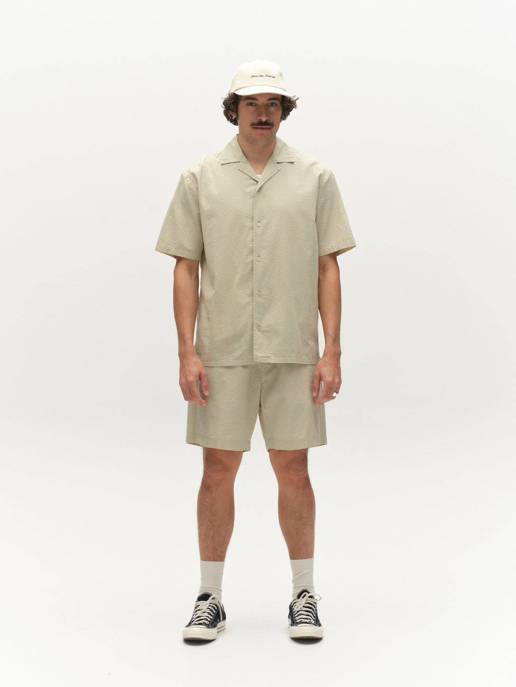 A man stands against a white background wearing a beige short-sleeve button-up shirt, Gabba GABtommi Ker Shorts in Alfa Green with an elasticated waistband, a matching beige cap, white socks, and black sneakers.
