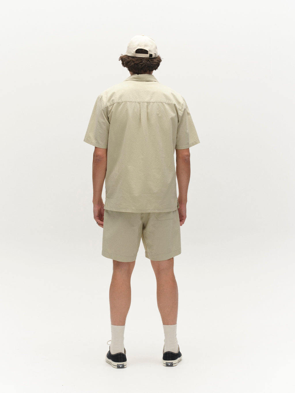 A person faces away from the camera, wearing a light beige short-sleeve shirt, Gabba GABtommi Ker Shorts in Alfa Green with an elasticated waistband, socks, black shoes, and a beige cap, against a plain white background.