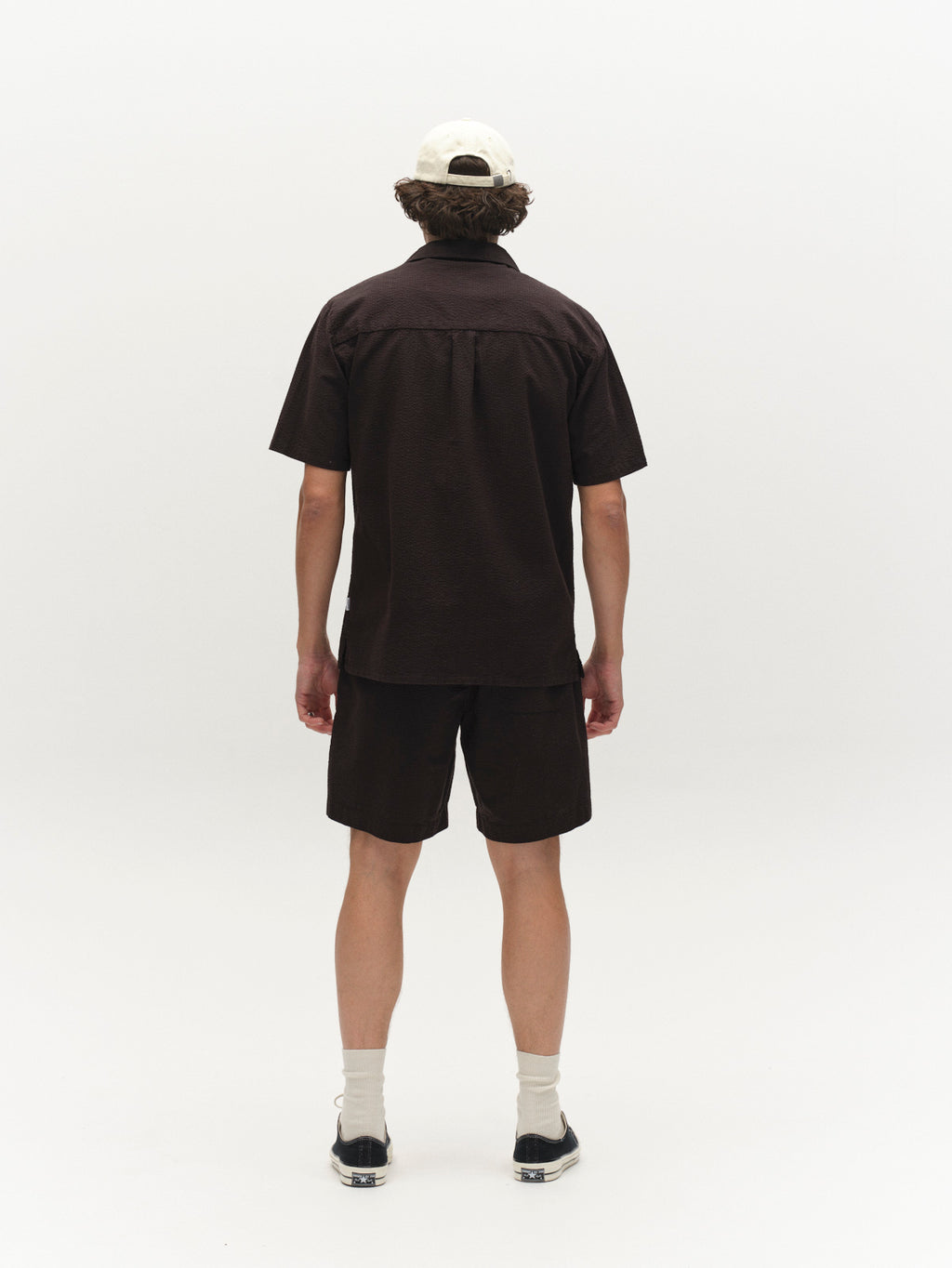 A person stands facing away from the camera, wearing a dark brown short-sleeve shirt, Gabba GABtommi Ker Shorts - Black with an elasticated waistband, a white cap, light socks, and dark sneakers against a plain white background.