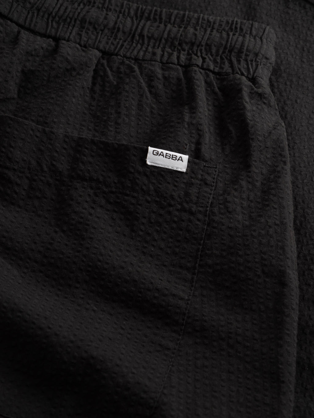 Close-up of black GABtommi Ker Shorts by Gabba, featuring a textured fabric, comfortable elastic waistband, regular fit, and a front pocket with a white GABBA tag.