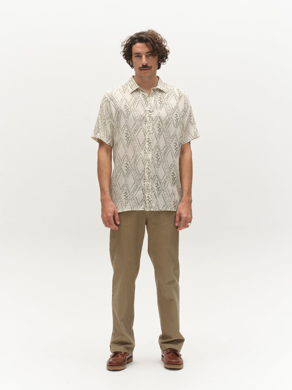 A man with wavy hair and a mustache stands facing forward, wearing the Gabba GABsven Reg Zag SS Shirt – Multi Pattern, tan pants, and brown shoes against a plain white background.