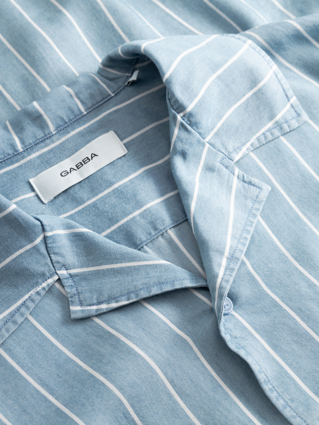 Close-up of the GABtimo Resort Stripes LS Shirt in light blue denim by Gabba, showing white vertical stripes, a folded collar, front pocket, and a Gabba label sewn inside the neckline.