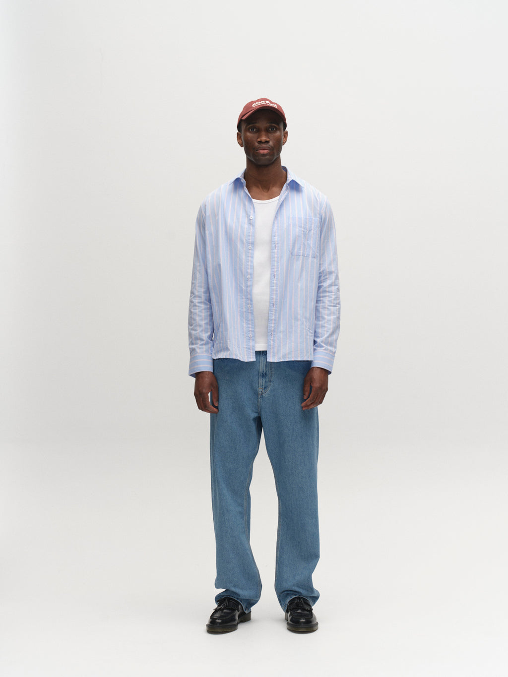 A man faces forward wearing the Gabba GABsven Reg Linea LS Shirt in rose stripes over a white tee, paired with blue jeans, black shoes, and a red cap against a plain white background.