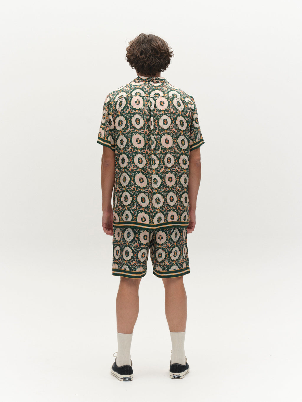 A person with curly hair faces away, wearing Gabba's GABtommi Oro Shorts in a green pattern, paired with a matching short-sleeve shirt, white socks, and black shoes against a plain white background.