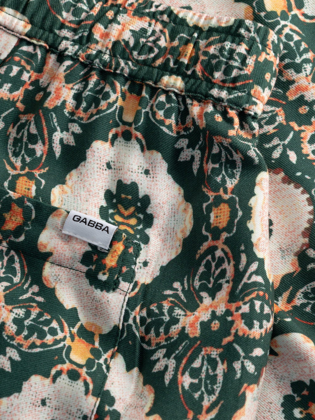 Close-up of the GABtommi Oro Shorts by Gabba, showcasing intricate green, orange, and cream floral patterned viscose fabric with a small white Gabba tag sewn near the seam.
