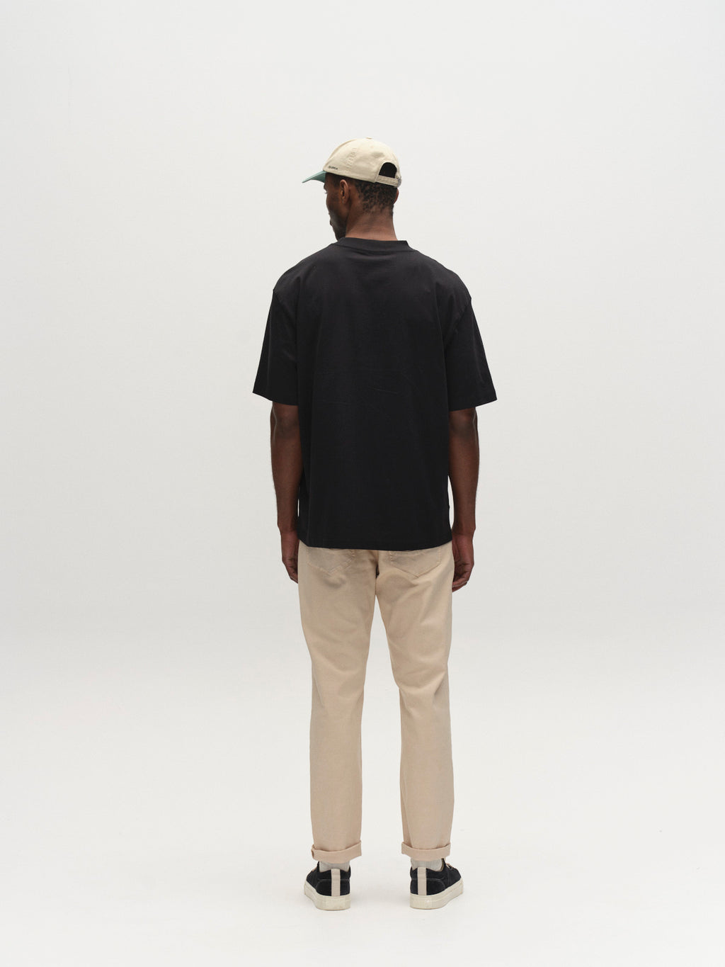A man stands with his back to the camera, wearing Gabba GABmarc 5poc Marlo - Birch jeans, a black t-shirt, black and white sneakers, and a light-colored cap against a plain white background.