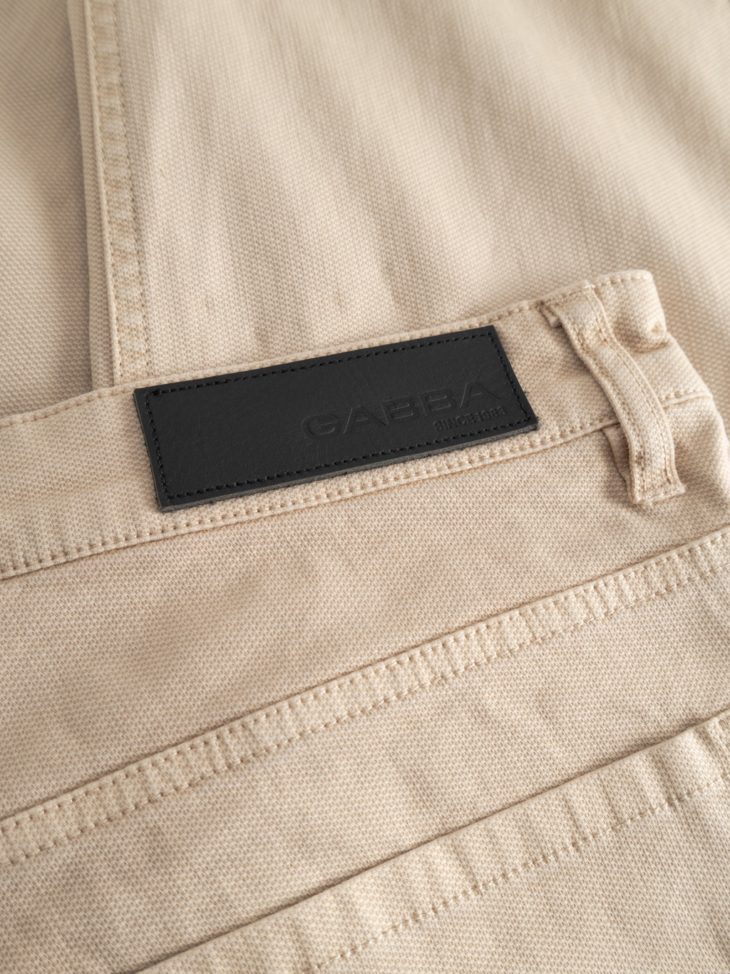 Close-up of beige textured fabric GABmarc 5poc Marlo - Birch jeans by Gabba, featuring a black rectangular waistband label. Visible stitching highlights their comfort and versatile style, ideal for everyday wear.