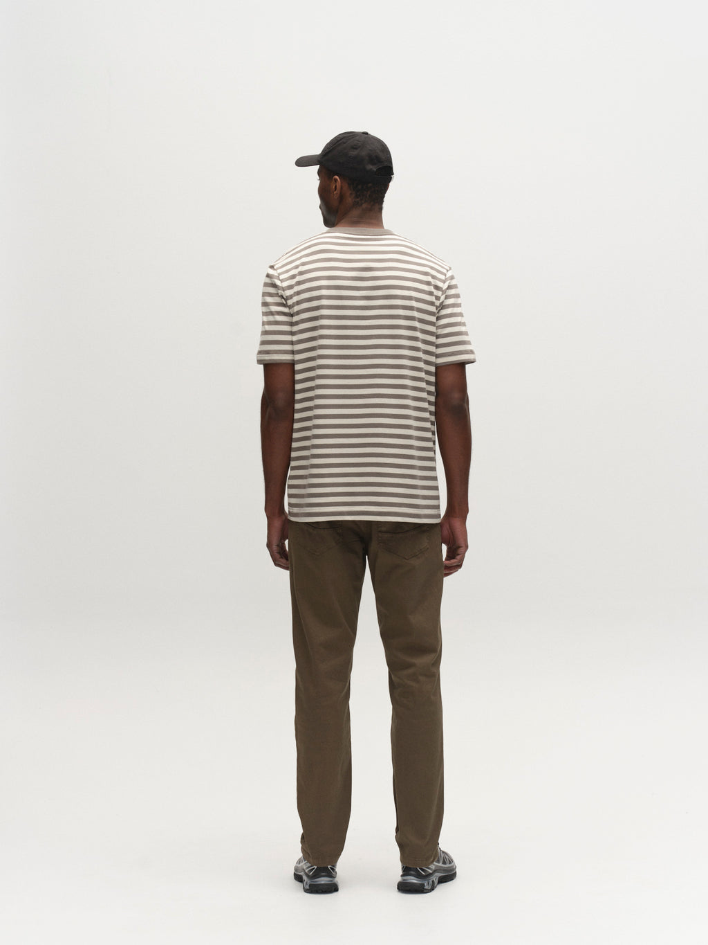 A man faces away from the camera, wearing a black cap, a beige and white striped tee, black sneakers, and Gabba GABmarc 5poc Marlo - Army jeans—ideal for everyday wear—posing against a plain white backdrop.