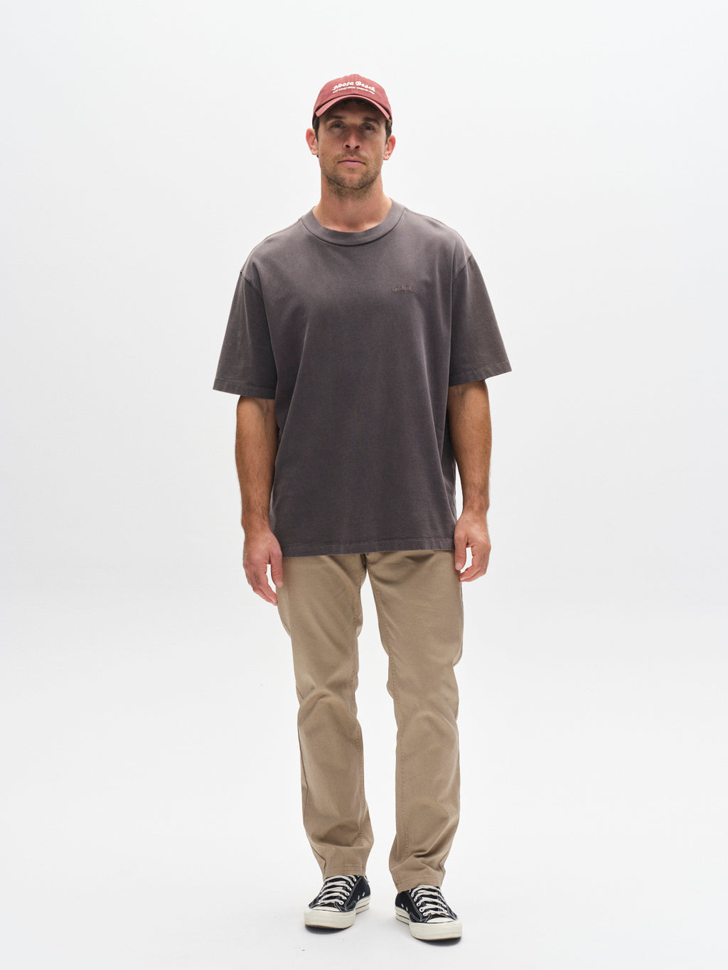 A man stands facing forward against a plain white background, wearing Gabba GABmarc 5poc Marlo - Coriander jeans, a faded dark gray t-shirt, black-and-white sneakers, and a red baseball cap.