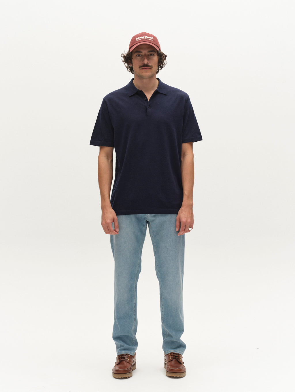 A man with a mustache stands against a white background, wearing a red cap, navy polo, Gabba GABmarc Hobok Jeans in mid blue denim, and brown shoes. He faces forward with his arms at his sides.