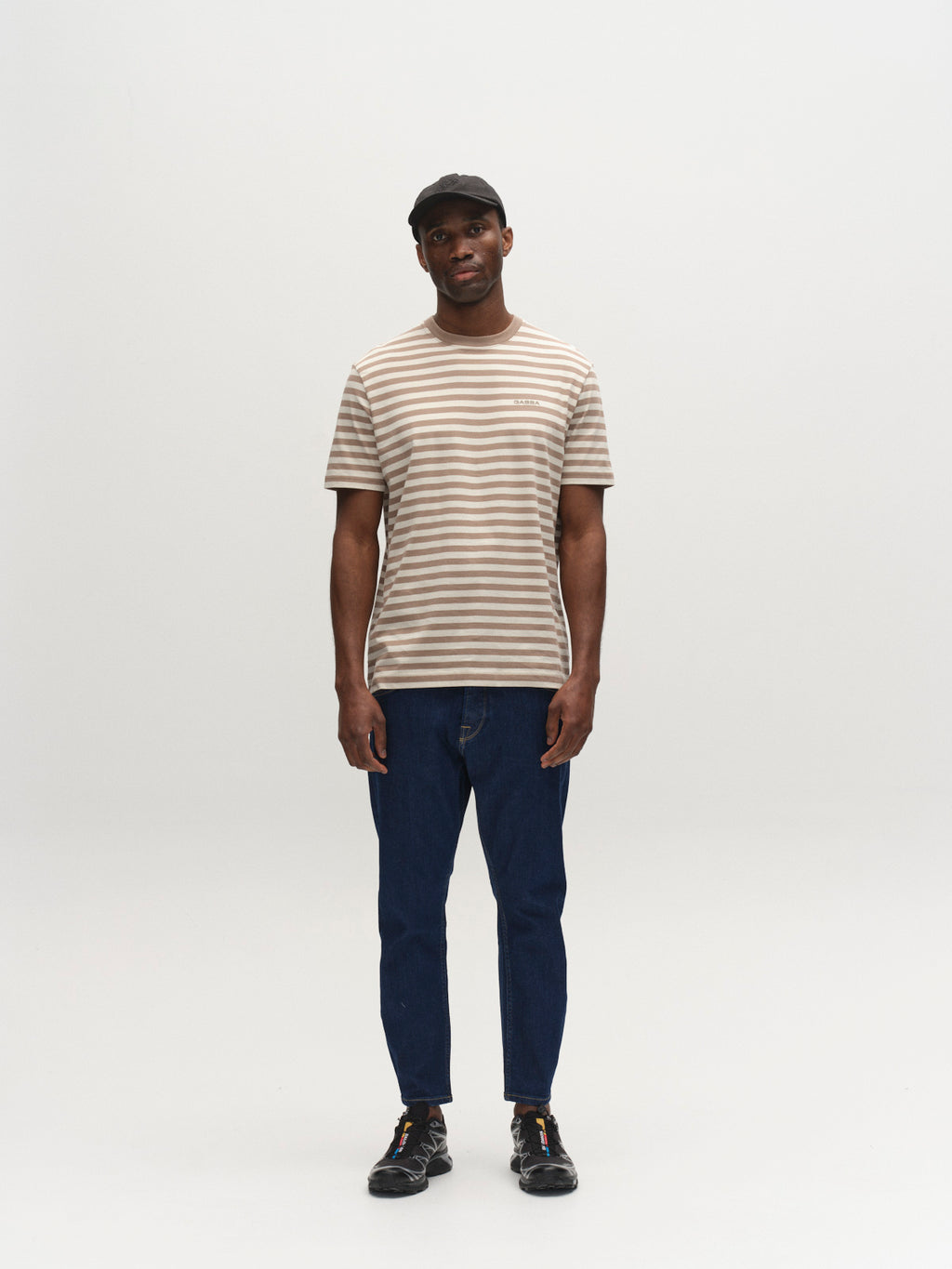 A man in a black cap, beige and white striped tee, Gabba GABalex SX Cropped Jeans in Dark Denim, and black sneakers stands facing forward against a plain white background.