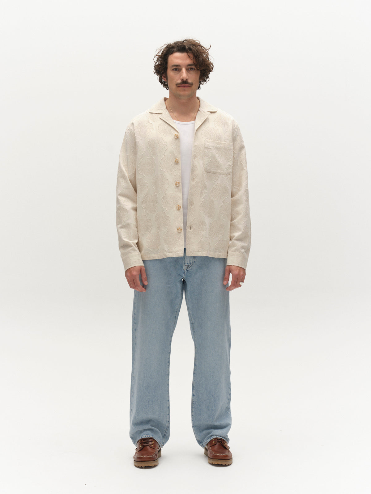 A man with curly hair and a mustache faces forward, wearing the Gabba GABaustin Reso Raw LS Overshirt in Lt. Sand, paired with light blue jeans and brown shoes, against a plain white background.