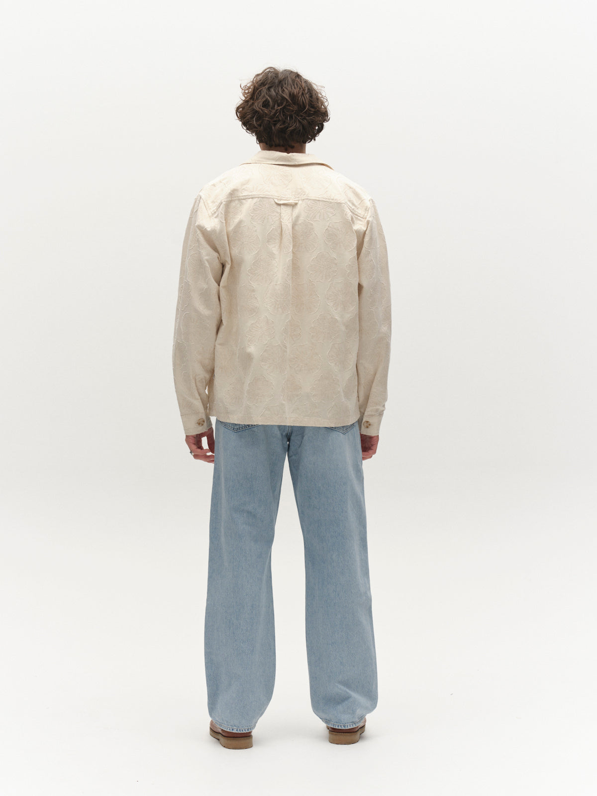 A person with curly hair stands facing away, wearing the Gabba GABaustin Reso Raw LS Overshirt in Lt. Sand and light blue wide-leg jeans against a plain white background.