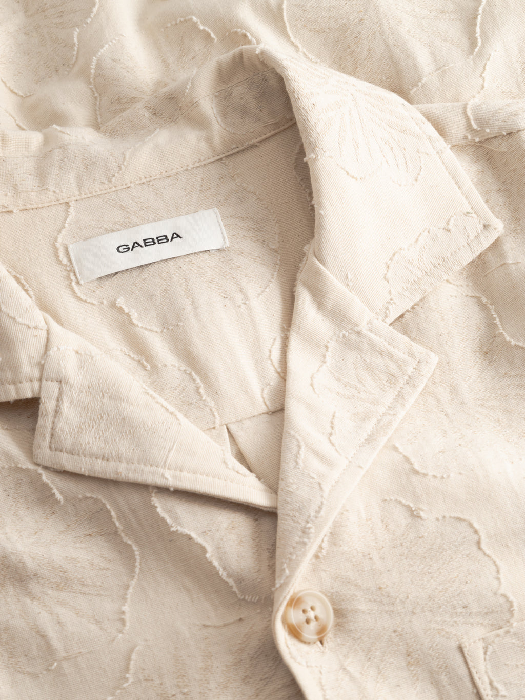 Close-up of a cream-colored GABaustin Reso Raw LS Overshirt by Gabba, featuring a textured cut-out floral pattern, notched lapel, button closure, and a white label with the Gabba brand name inside the collar.