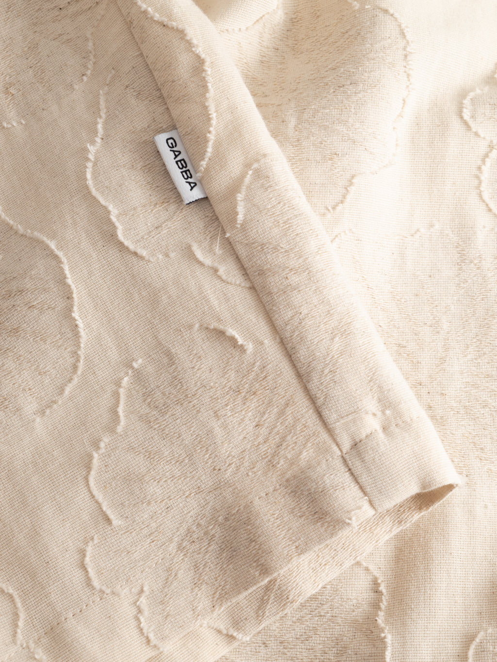 A close-up of the Gabba GABaustin Reso Raw LS Overshirt in Lt. Sand shows soft, jacquard-woven cotton with a subtle cut-out floral pattern, visible white CABBA label, and gentle fabric folds and stitching.