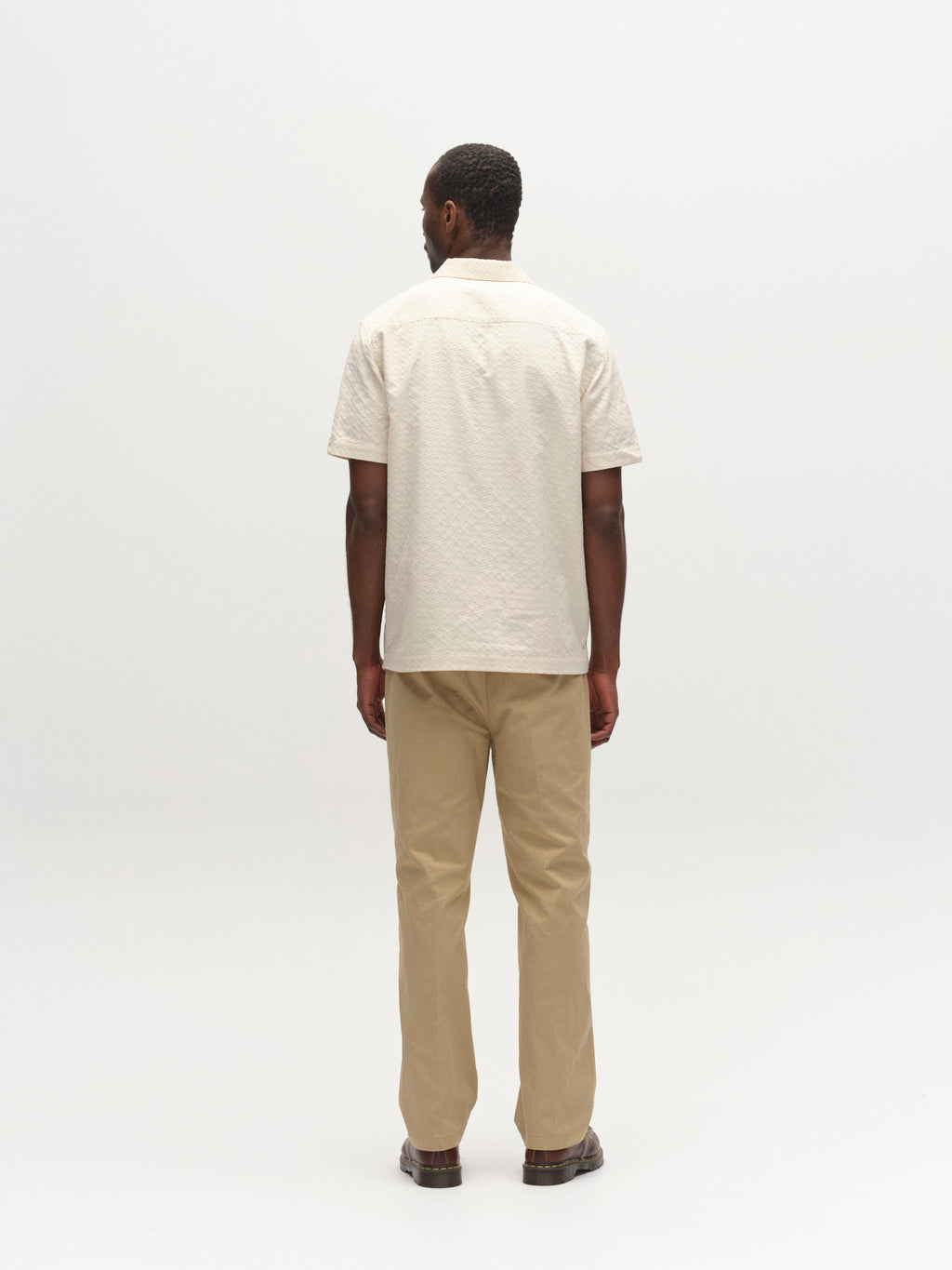 A man stands with his back to the camera against a plain white background, wearing the Gabba GABtimo Resort Neal SS Shirt in Birch, along with tan pants and brown shoes.