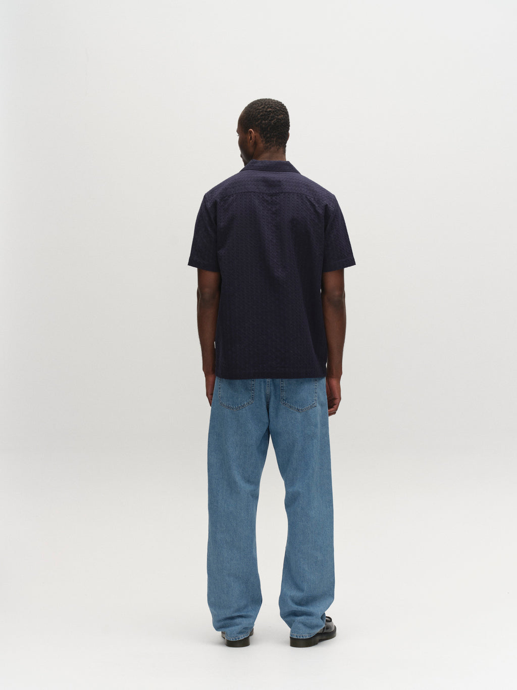 A man stands against a plain white background, facing away from the camera. He wears the Gabba GABtimo Resort Neal SS Shirt in Navy, paired with loose-fitting blue jeans and dark shoes, his arms resting at his sides.