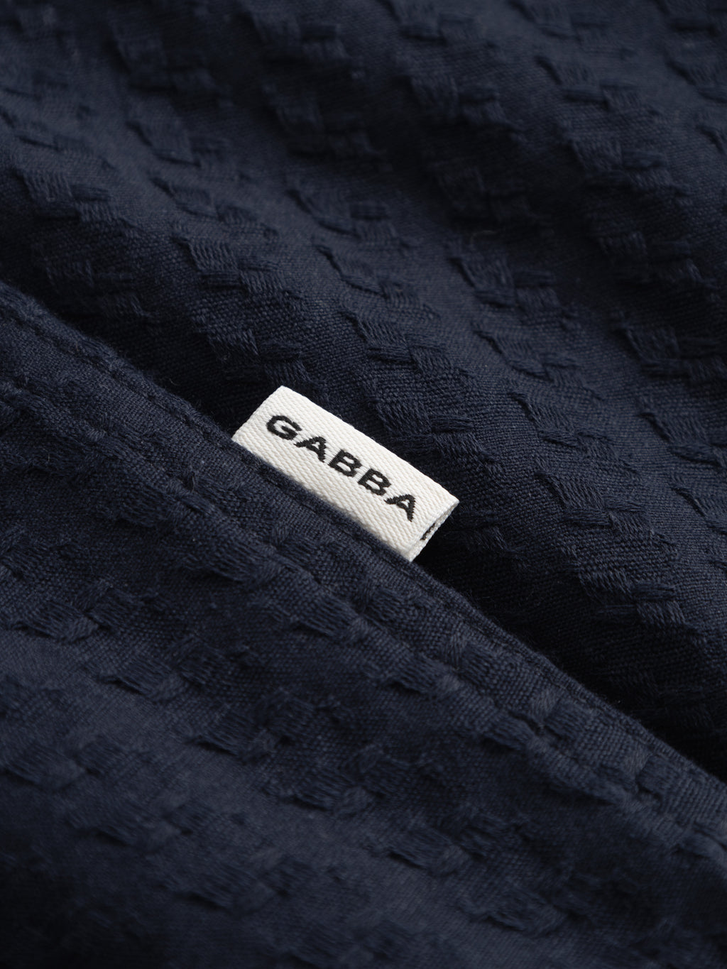 A close-up of textured dark navy fabric from the Gabba GABtimo Resort Neal SS Shirt, featuring a small white tag with 