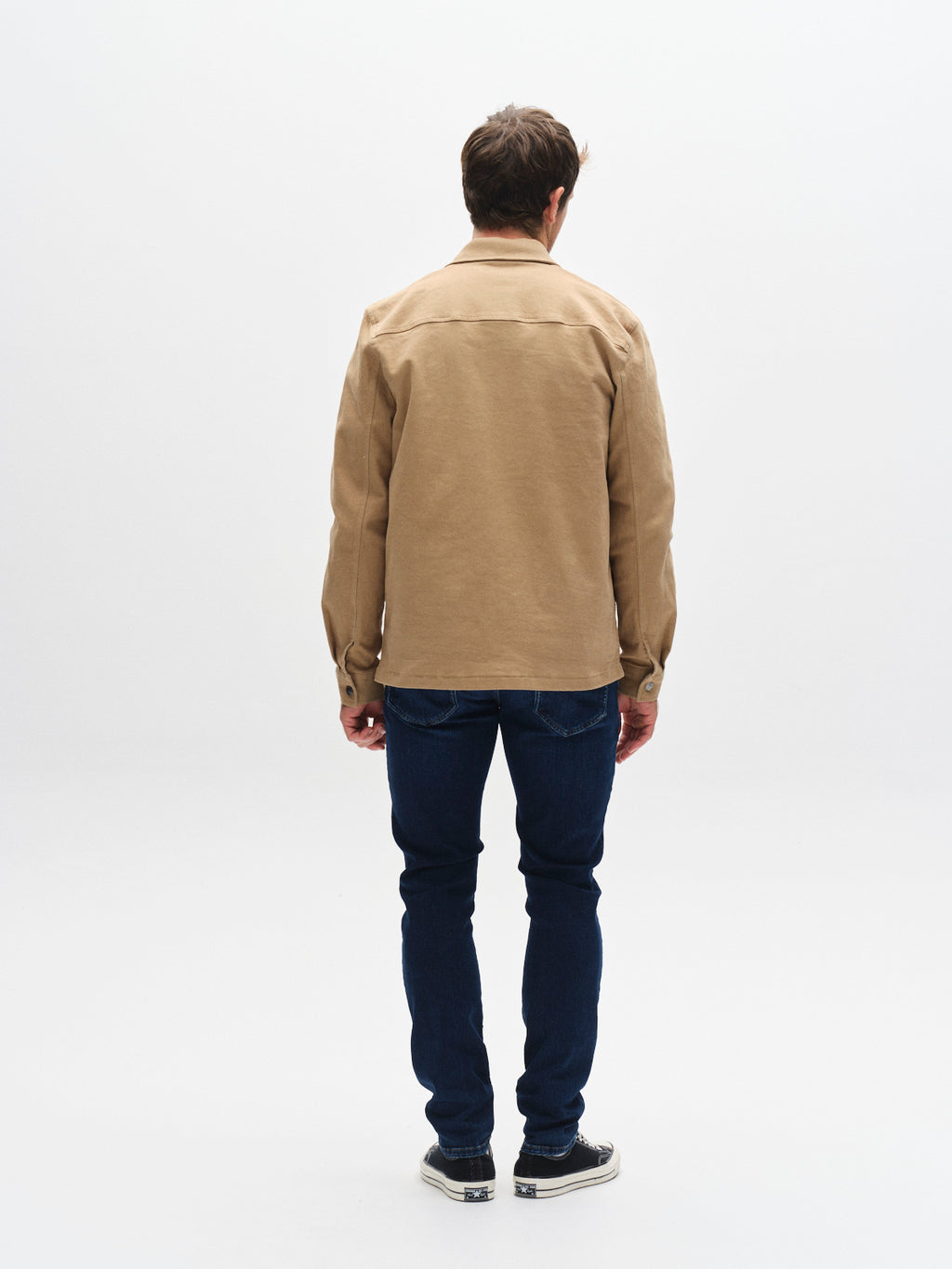 A man stands facing away from the camera, wearing a tan jacket, black and white sneakers, and Gabba GABjones Dhera Jeans in mid blue denim—crafted from superstretch fabric—against a plain white background; a modern wardrobe staple.