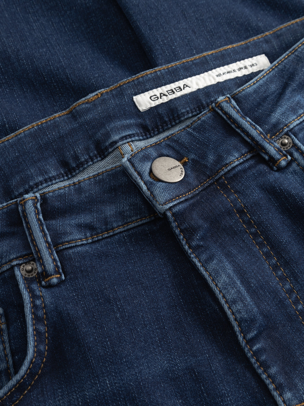 Close-up of Gabba GABjones Dhera Jeans in mid blue superstretch denim features waistband, metal button, belt loops, a white Gabba label, and classic orange stitching—a modern slim-fit essential.