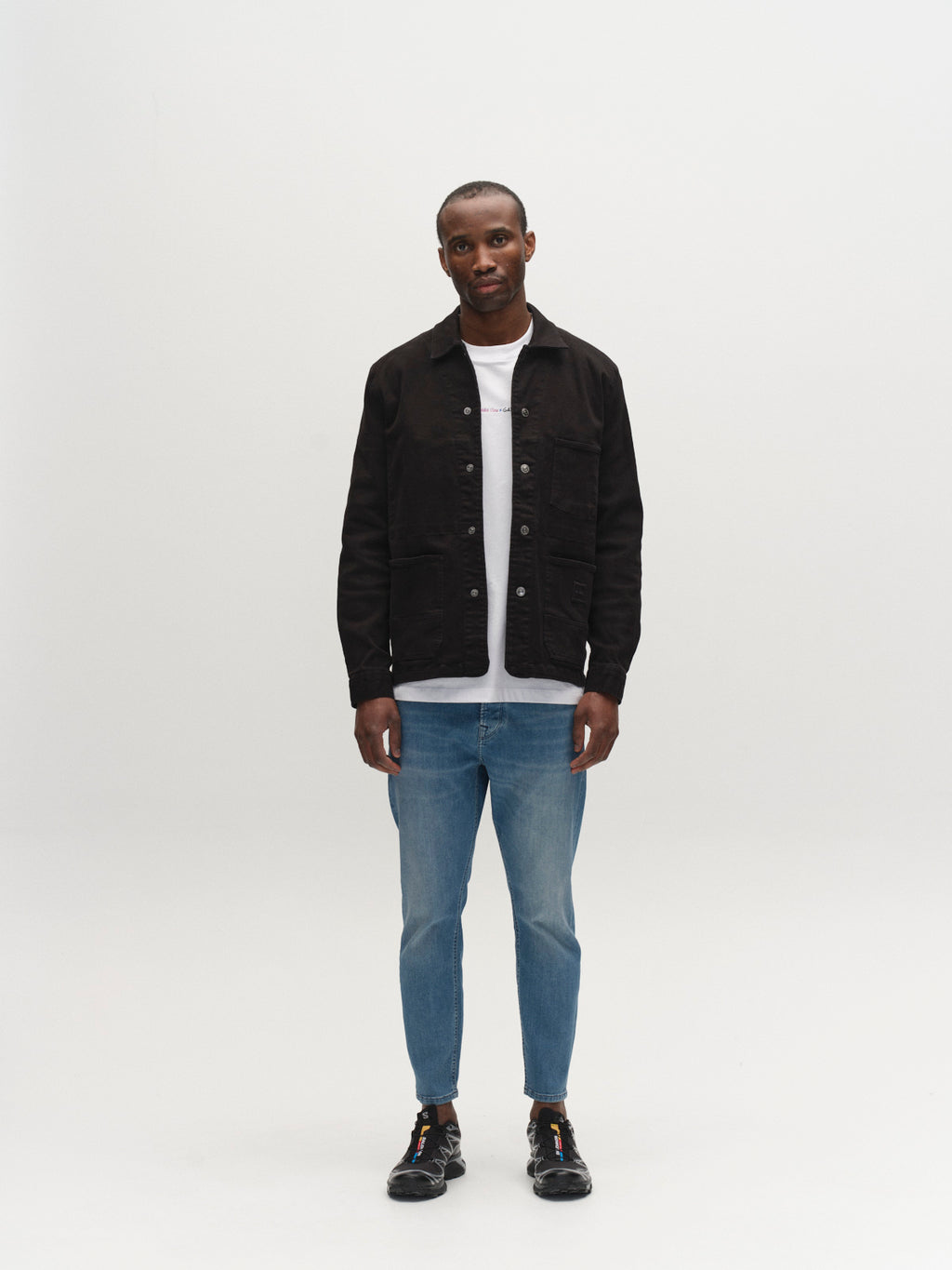 A man stands facing forward against a white background, wearing Gabba GABalex Dhera Cropped Jeans in Lt. Blue Denim, a black jacket over a white t-shirt, and black shoes. His arms rest at his sides and he has a neutral expression.