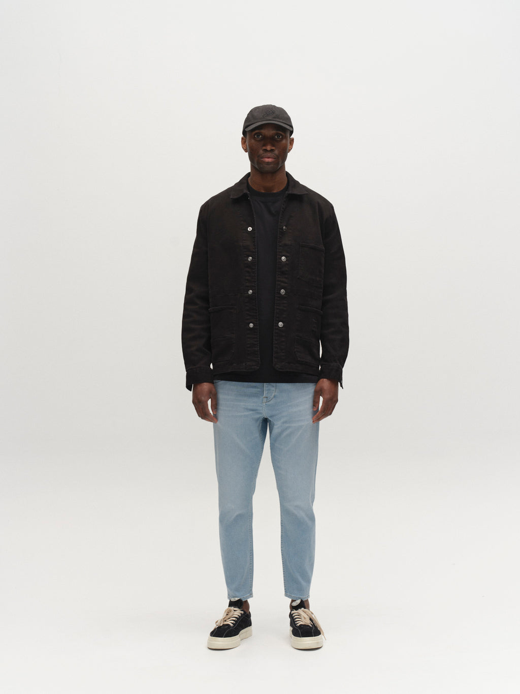 A person stands facing forward against a plain white background, wearing a black cap, black button-up jacket, black shirt, GABalex Dynamic Cropped Jeans - Lt. Blue Denim by Gabba, and black and white sneakers.