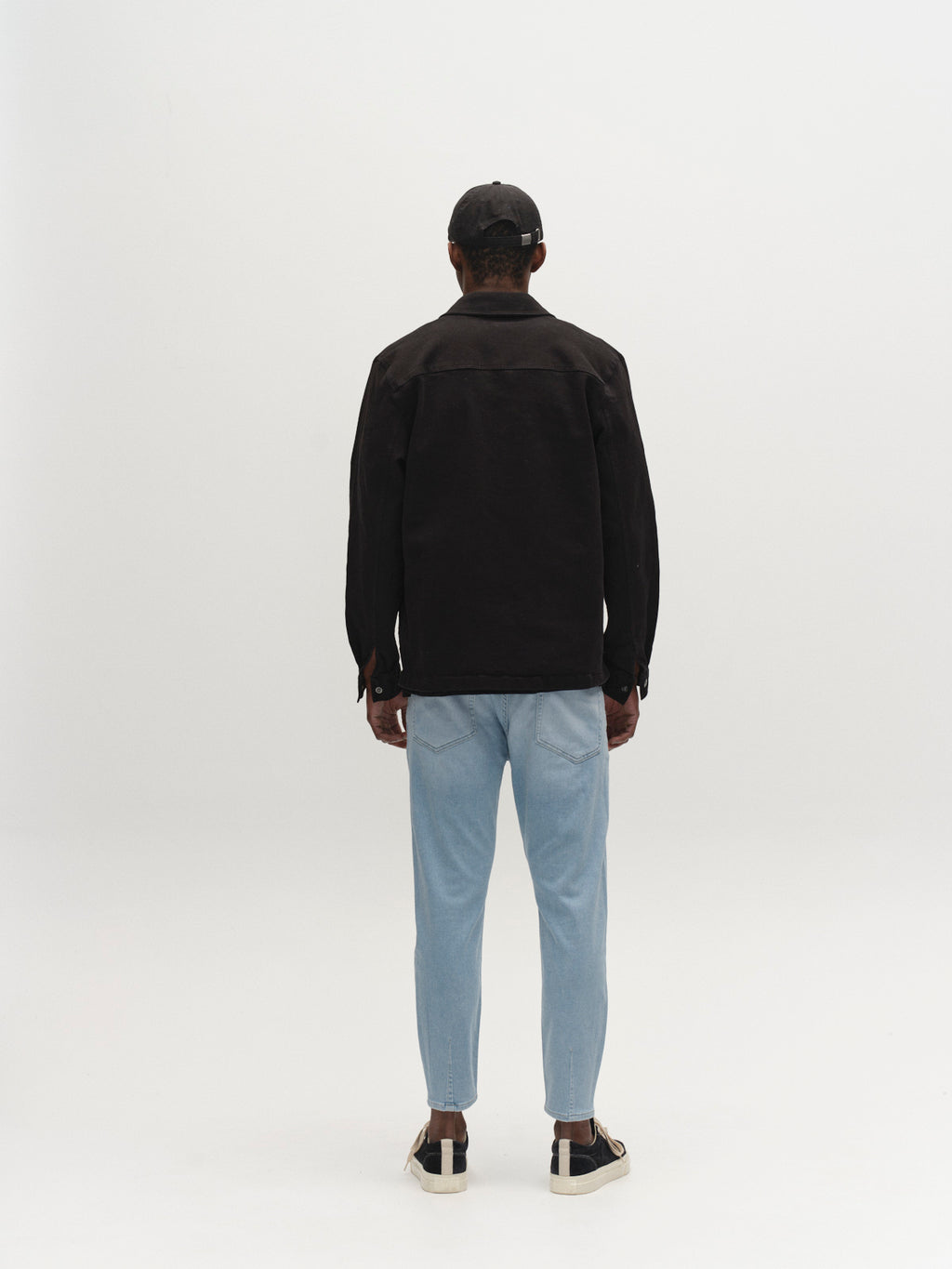 A person faces away from the camera, wearing Gabba GABalex Dynamic Cropped Jeans in Lt. Blue Denim with a black jacket, black cap, and black-and-white sneakers against a plain white background.