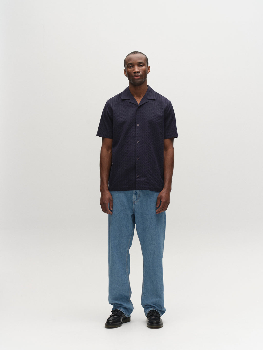 A man faces forward against a plain white background, wearing a short-sleeved dark shirt, black shoes, and Gabba GABnoah Brook Jeans in denim wash with a relaxed silhouette and neutral expression.