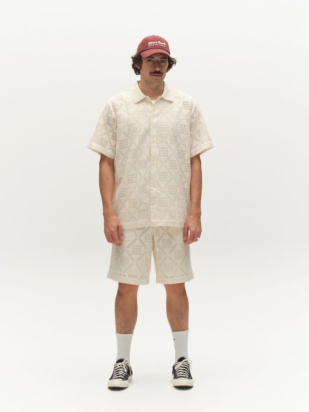 A man wearing Gabba’s GABjoli Ted Croch Shorts GRS in Birch, a cream relaxed-fit crochet short-sleeve shirt, red cap, white socks, and black sneakers stands against a plain white background.