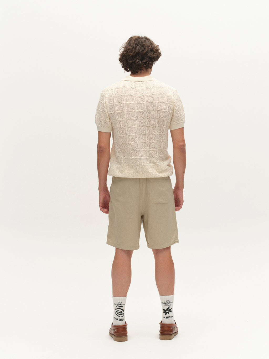 A person with curly hair faces away, wearing the Gabba GABbenson Fiwi SS - Birch short-sleeve knit polo, beige shorts, white socks with black designs, and brown shoes, set against a plain white background.