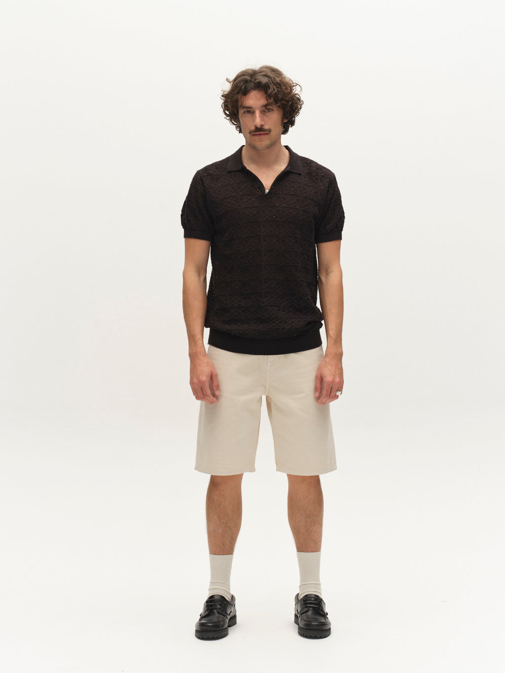 A man with curly hair and a mustache stands against a plain white background, wearing the Gabba GABbenson Fiwi SS - Black polo, beige knee-length shorts, cream socks, and black loafers.