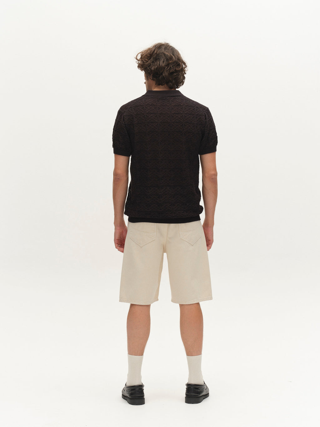 A person with wavy brown hair stands facing away, wearing the Gabba GABbenson Fiwi SS polo in black, beige knee-length shorts, light socks, and black shoes against a plain white background.