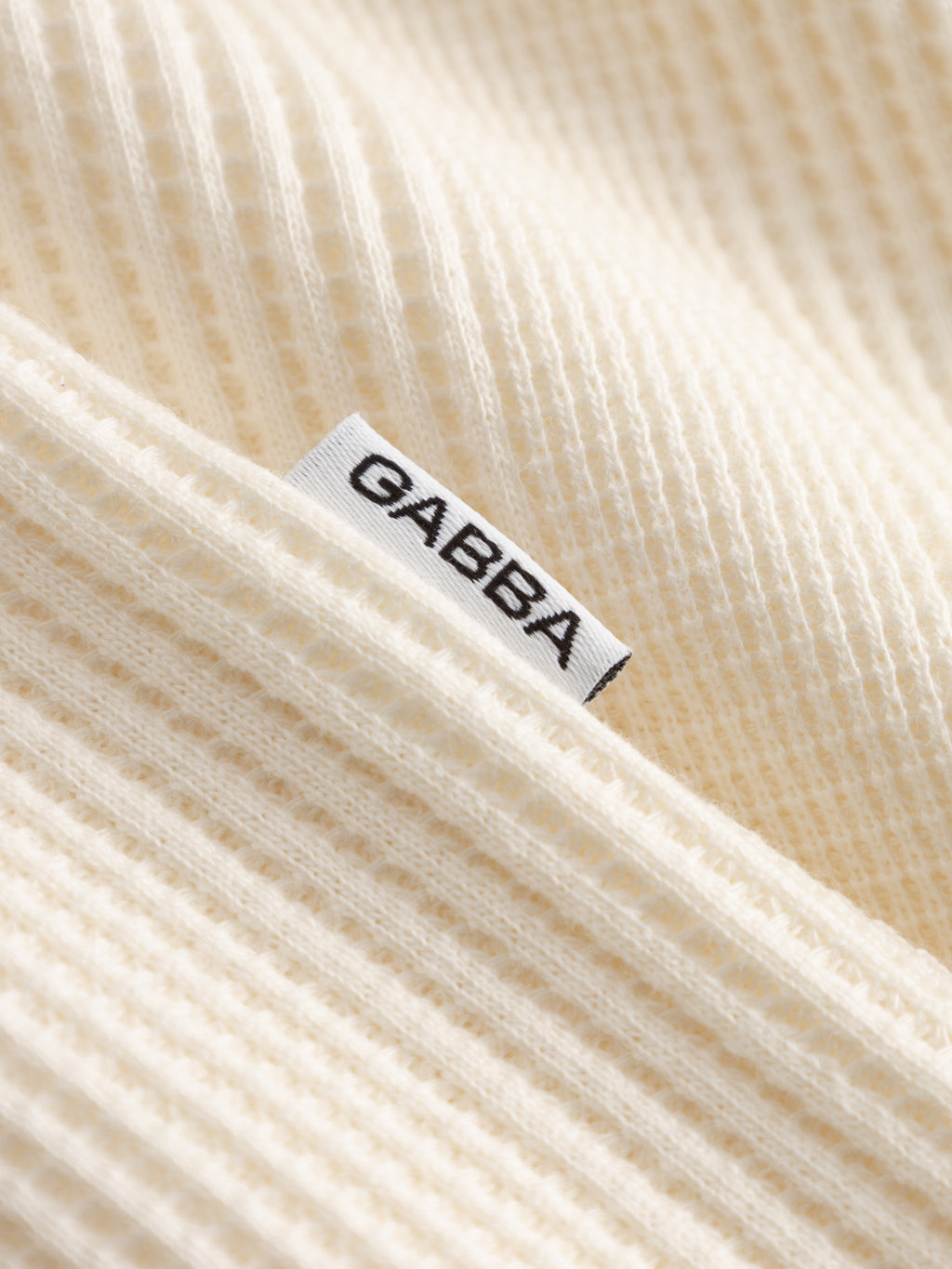 A close-up of the GABvalente Wup SS T-Shirt by Gabba in Bone White, featuring soft, ribbed fabric and a white tag with GABBA in black uppercase letters, evoking the cozy feel of a waffle-textured shirt.