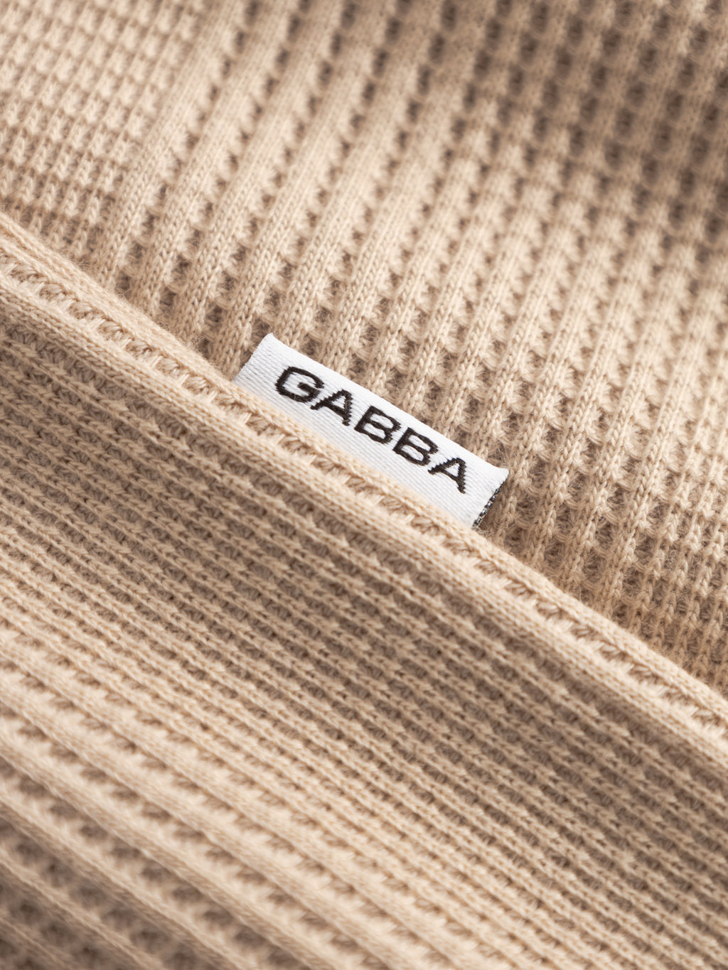 Close-up of beige waffle-textured fabric from the GABvalente Wup SS T-Shirt - Humus by Gabba, featuring a small white tag stitched into the seam with the brand name in bold black letters.