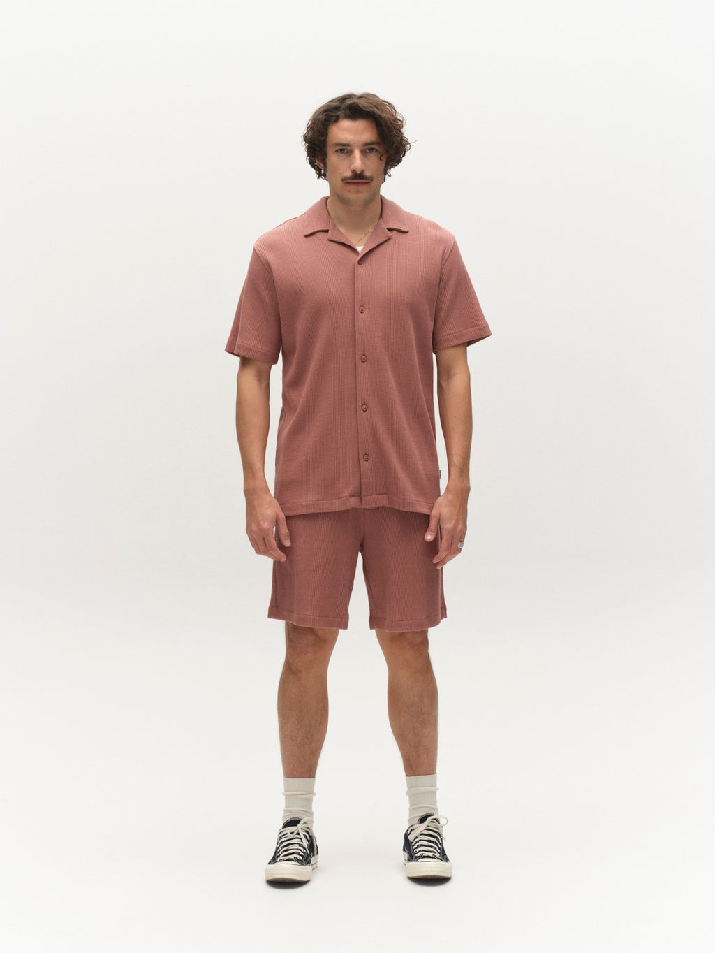 A man with a mustache stands facing forward in a relaxed fit Gabba GABvalente Wup SS T-Shirt in Wood Pink, paired with shorts, white socks, and black sneakers against a plain white background.