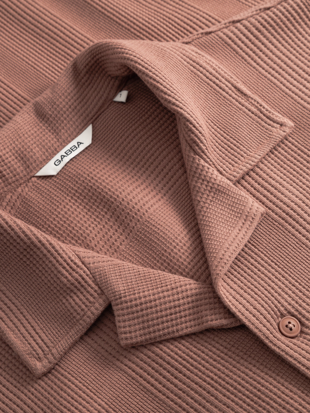 A close-up of the Gabba GABvalente Wup SS T-Shirt in wood pink shows its waffle texture, button detail, and open resort collar. Inside the collar is a white tag with 