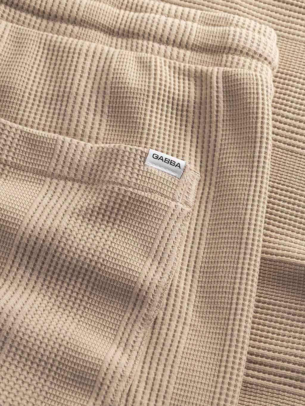 A close-up of GABtommi Wup Shorts - Humus by Gabba shows beige waffle-textured fabric with a folded finish and a small white tag labeled 