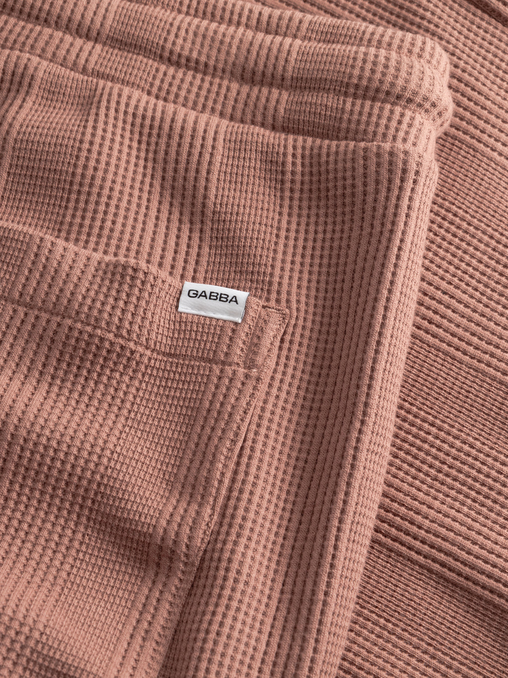 A close-up of Gabba’s GABtommi Wup Shorts in Wood Pink shows the waffle texture and comfy waistband, with a folded section revealing a sewn-in white label featuring the brand's name.