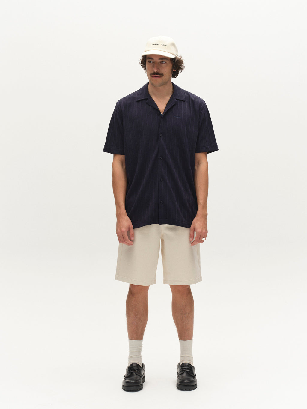 A man stands against a plain white background wearing a Gabba GABvalente Armor SS T-Shirt in navy, a cream baseball cap, beige shorts, beige crew socks, and black loafers.