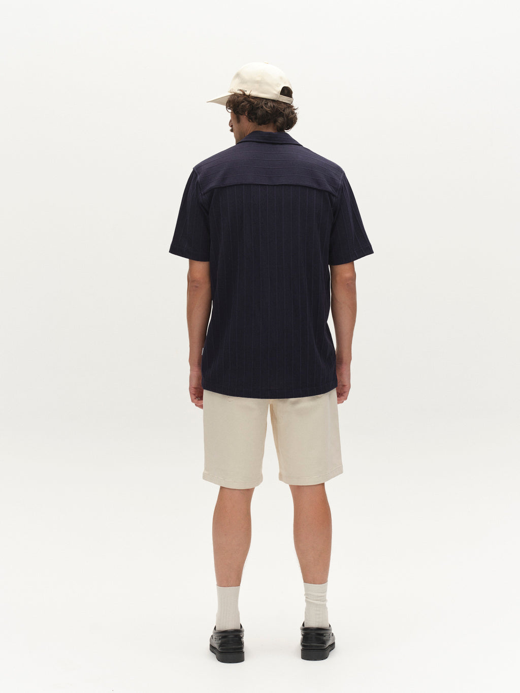 A person stands against a plain white background, facing away and wearing the Gabba GABvalente Armor SS T-Shirt in navy, a cream-colored cap, cream shorts, light socks, and black shoes.