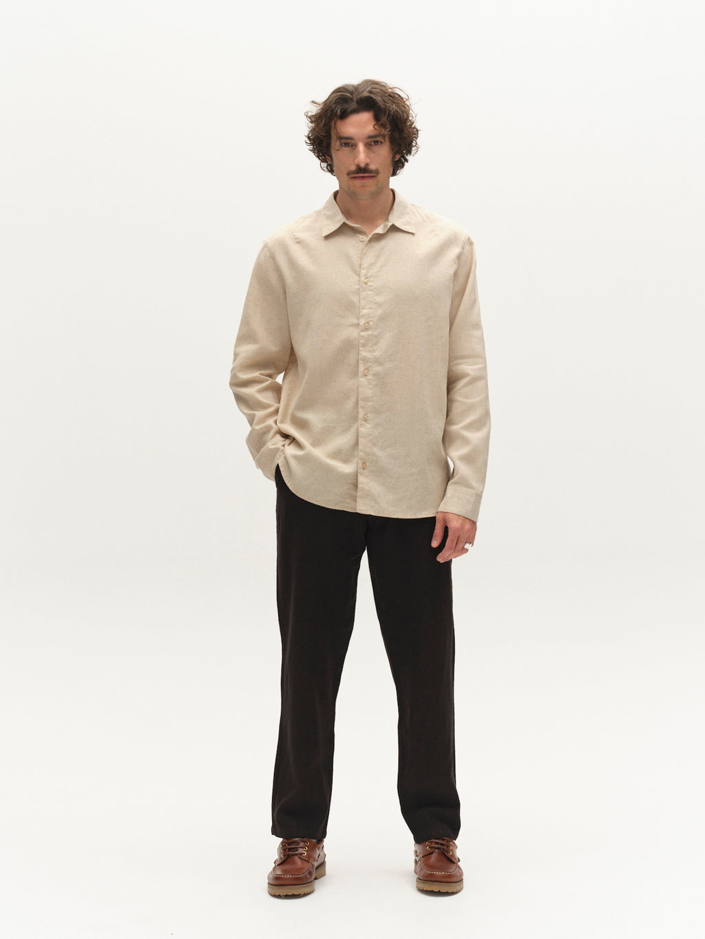 A man with curly hair and a mustache stands against a white background, wearing a beige button-up shirt, brown shoes, and Gabba’s GABnapoli 19 Lino Pant in Meteorite. One hand is in his pocket.