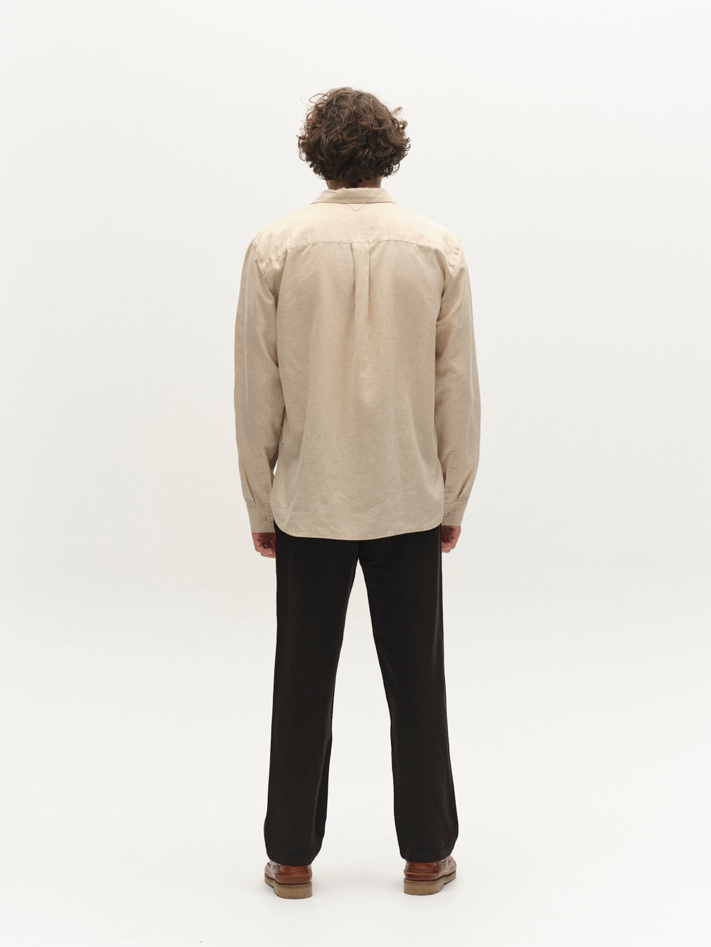A person with curly hair stands facing away from the camera, wearing a beige long-sleeve shirt and Gabba's GABnapoli 19 Lino Pant - Meteorite, paired with brown shoes, against a plain white background.